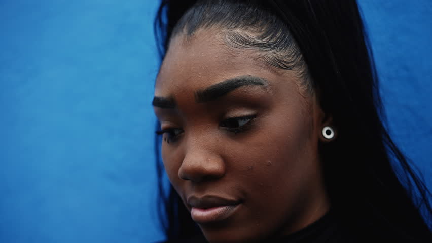 One pensive worried black adolescent girl with thoughtful expression, close-up face tracking shot in deep mental soulful reflection standing urban street gazing down