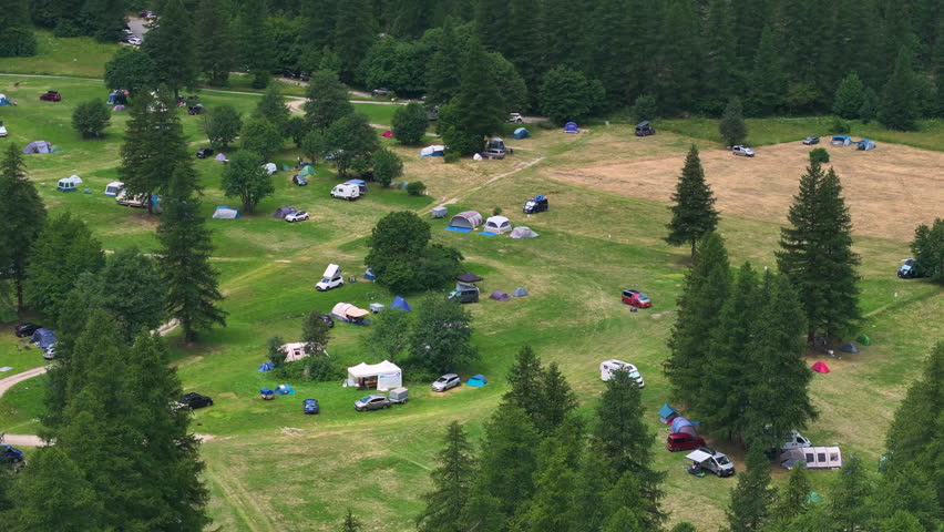 AERIAL: Campground nestled in a green alpine valley surrounded by dense forest. Tents and vehicles are scattered across the grassy clearing in the middle of mountainous alpine nature in French Alps.