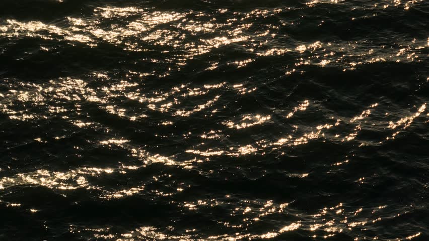 Sunset light dances on the sea’s surface, with waves creating reflective patterns in slow motion. The sunlight sparkles off the water, enhancing the mesmerizing texture of the ocean.

