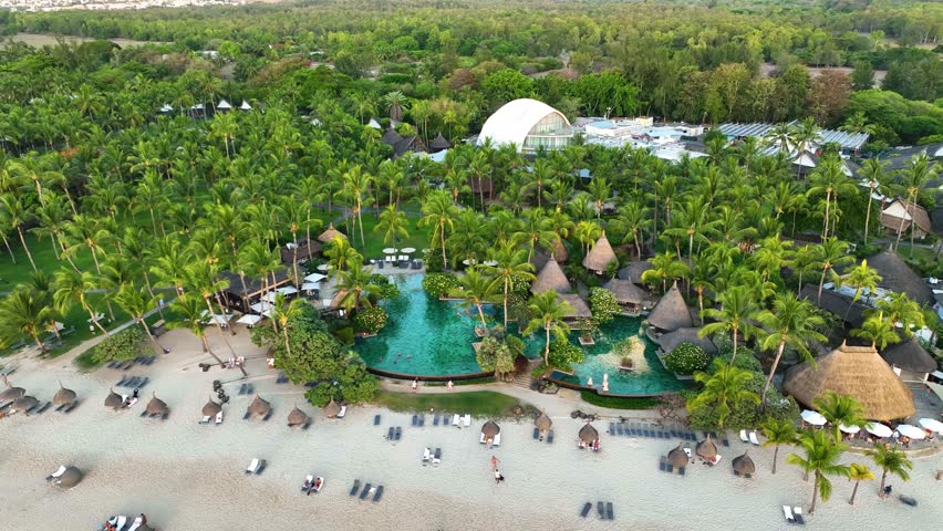 Overhead view of a beachfront resort in Mauritius with a lush palm tree garden, thatched roof villas, turquoise swimming pools, and sunbeds on the sandy shore
