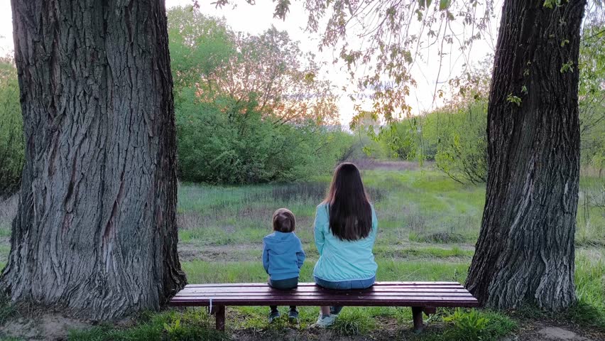 Back of mother and child sitting at park bench together. Parent and son enjoying nature in sunny day. Mother and little son resting on bench near park, family weekend in park, relax.