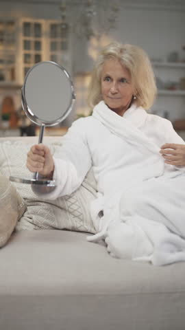 Pretty mature woman admiring her face in mirror, better aging and natural beauty . Stay attractive in old age, beautiful grey-haired lady relaxing after beauty routine and home spa, relax and be calm