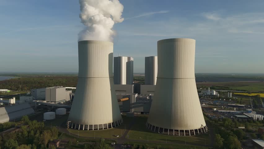 Drone flies towards the front of the Lippendorf power plant cooling towers near Leipzig, Germany. Massive infrastructure with steam and clear blue sky on a spring day.