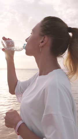 Quench thirst after morning run or workout on seashore, woman drinking water. Slender young adult lady sipping fresh water from plastic bottle, admiring sunrise or sunset above ocean, training outdoor