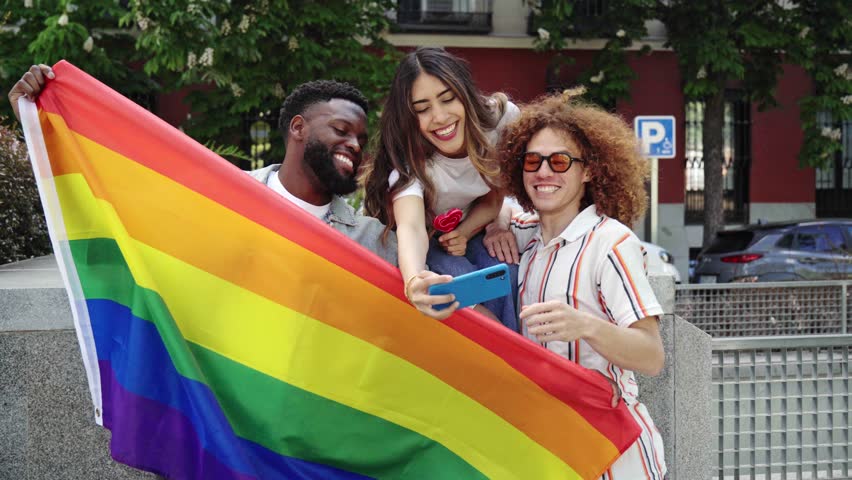 Happy multi-ethnic friends are taking selfies with a smartphone, holding a rainbow flag, symbolizing lgbtq+ pride and inclusivity, in an urban setting