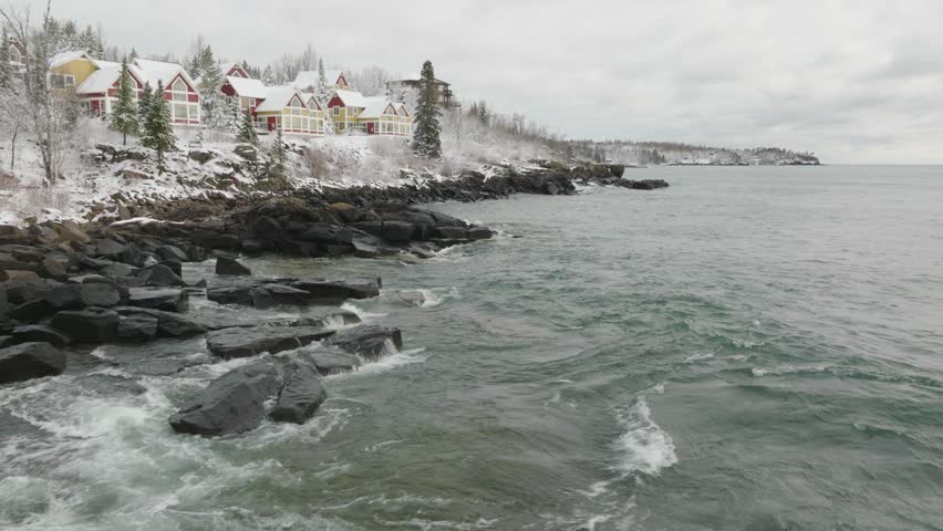 Waves Splashing On Rocky Shore Of Lake Superior In Beaver Bay, Minnesota. Snow-covered Cottages On Rugged Coastline. drone pullback shot