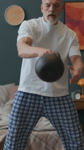 Vertical shot of athletic senior man in homewear standing in his bedroom and performing kettlebell swings during daily workout routine