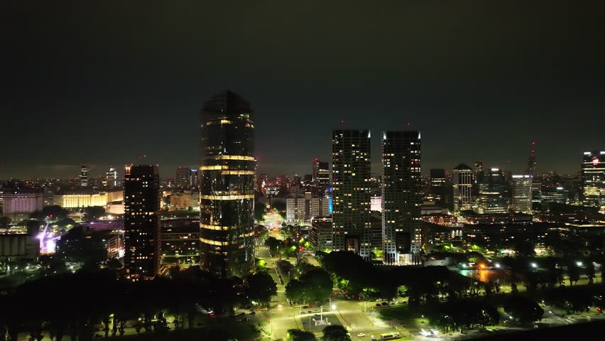 Aerial night view of downtown Buenos Aires with illuminated buildings, cars on city streets, and trees, extending toward the glowing city horizon.