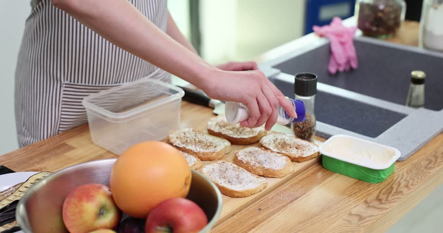 Woman in apron sprinkles salt over toasts topped with cream cheese while plate with grilled eggplants and bowl with fruits stand nearby in kitchen
