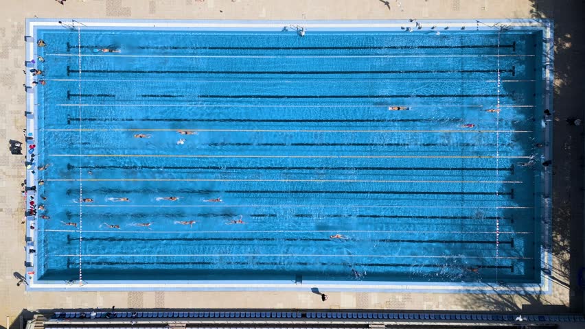Top-down aerial view of swimmers training in an outdoor swimming pool with clear blue water and visible poolside activity