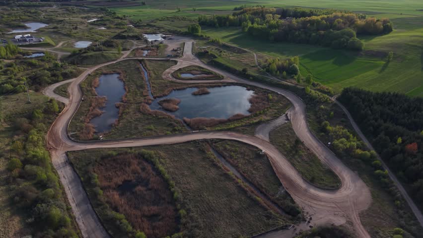 Elevated view of winding dirt tracks near ponds and green forested areas under soft spring light, showcasing natural and rural land use patterns.