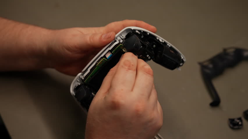 Close-up of technician hands examining internal components of disassembled video game controller, highlighting intricate joystick mechanisms and circuit board details. Shooting in slow motion.