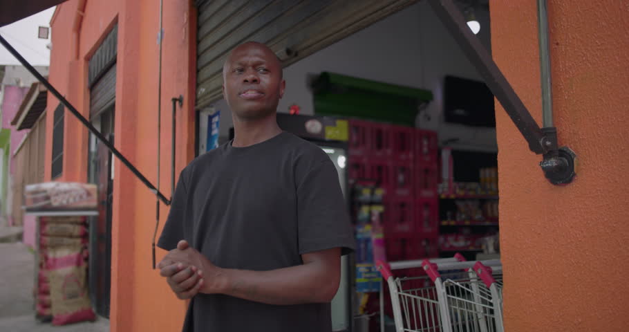 Happy African American shop owner smiling outside neighborhood grocery store with open gate, standing confidently in front of orange wall at start of day