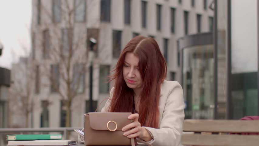 In a cozy cafe with greenery, a woman with long red hair sits at a table, taking notes in her notepad as she works