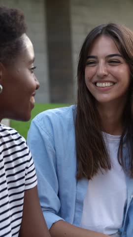 Vertical HD video of happy millennial female friends talking outdoor. Multiracial young girls having fun together laughing while sitting on stairs in city street. Youth lifestyle concept