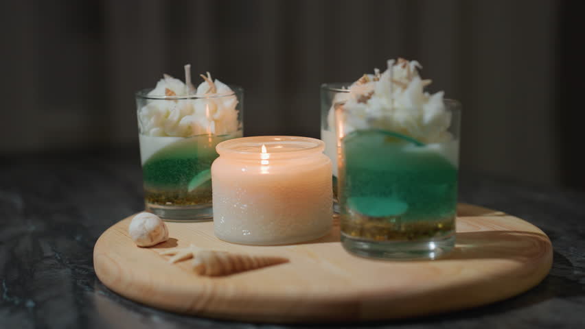Round view of beautifully arranged candle containers on wooden tray, one candle lighted casting soft glow and gentle reflection on nearby unlit decorative candles surrounded by elegant cream swirls