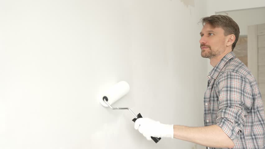 Professional male painter rolling white paint on apartment wall, transforming interior space with steady strokes during comprehensive home renovation