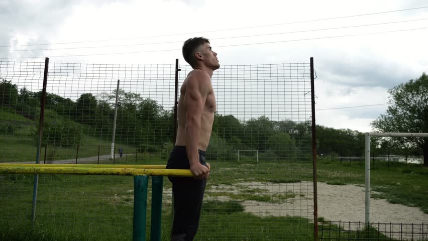 Strong man trains abdominal muscles raising legs on parallel bars at sports ground. Young shirtless sportsman exercising at nature. Sporty athlete working out at nature. Concept of sportive lifestyle