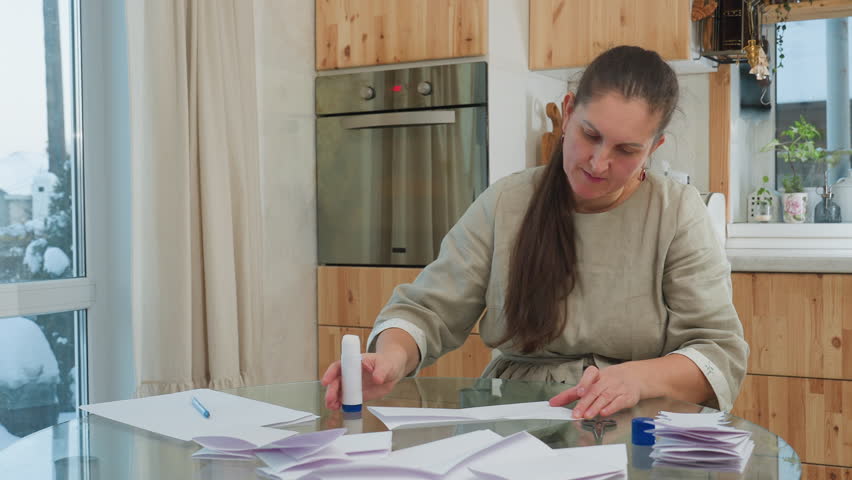 Woman applying glue to paper while sitting at table with scissors, folded paper, and other crafting supplies in background