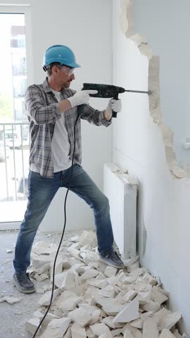 Man construction worker demolishing a wall with a hammer drill in protective gear, creating construction debris on the surrounding floor surface. Renovation concept