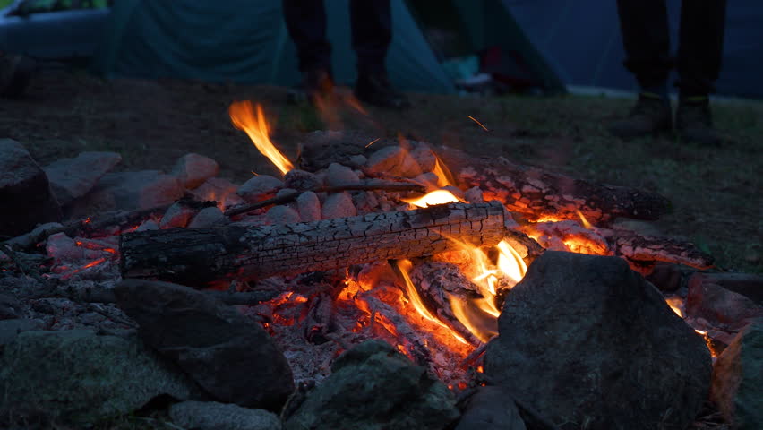 CLOSE UP: Crackling campfire in early evening with glowing embers and charred logs surrounded by stones. Two unrecognizable people stand by the fireplace positioned in the middle of a camping ground.