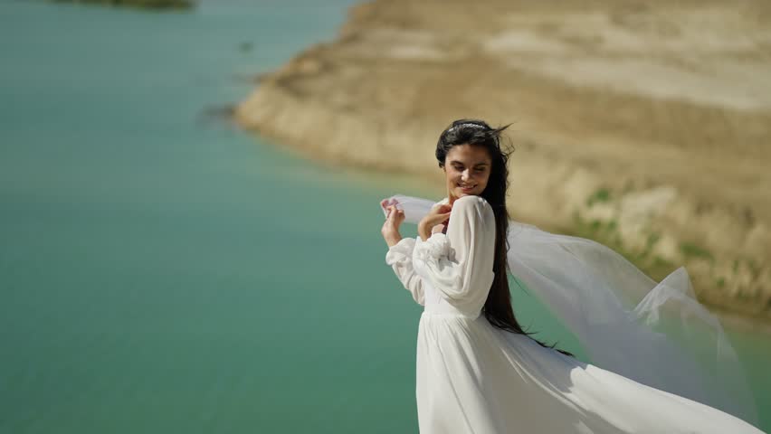 Woman in white dress near turquoise water on windy day, Elegant lady posing with flowing fabric by scenic cliffside lake view