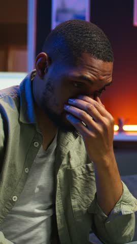 Vertical Video African american guy being overworked with headache at modern loft, working remotely under stress and pressure from his boss. Young person frustrated and worried, struggling with tasks