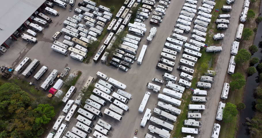 Large parking lot of local dealer with many new RV trailers parked for sale. Distribution of manufactured camping vehicles.