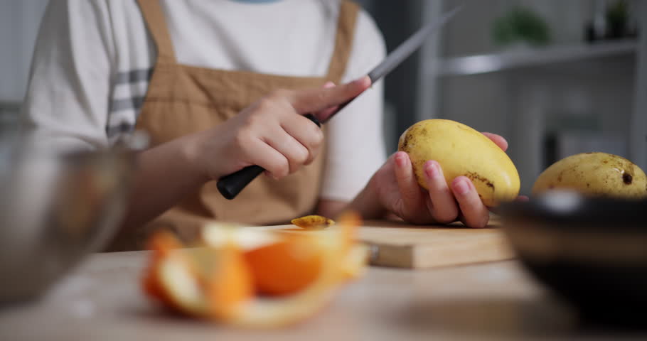 Handheld selective focus shot, Woman's hand using knife to peel ripe mango on wooden table. Preparing sweet food