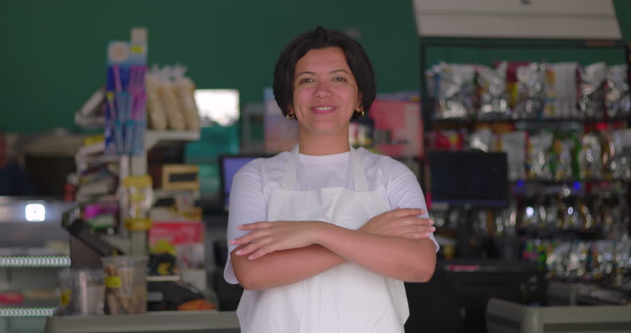 Smiling Latina woman wearing apron at supermarket checkout, casual confident portrait with colorful background, cheerful indoor retail setting