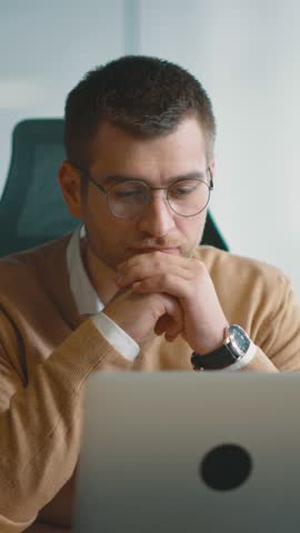 Smart looking focused businessman working on a laptop and thinking. Millennial managing family budget, handling financial paperwork, and tracking expenditures on a laptop at a modern open space office