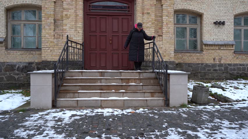 Elderly woman slowly descends the slippery steps of the stairs. Holding on to the railing of the porch of the house. A grandmother goes for a walk in winter. Street outdoor
