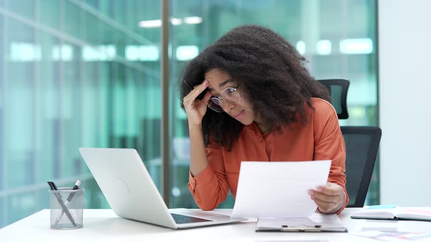 Frustrated african american businesswoman having difficulty with paperwork using laptop sitting at desk at workplace in a business office. Confused female worker having problems working with documents