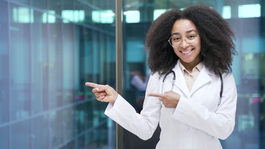 Happy young african american female doctor pointing with fingers at empty space for writing text standing in hospital clinic office, looks at camera. Template for advertising. Copy space for promotion