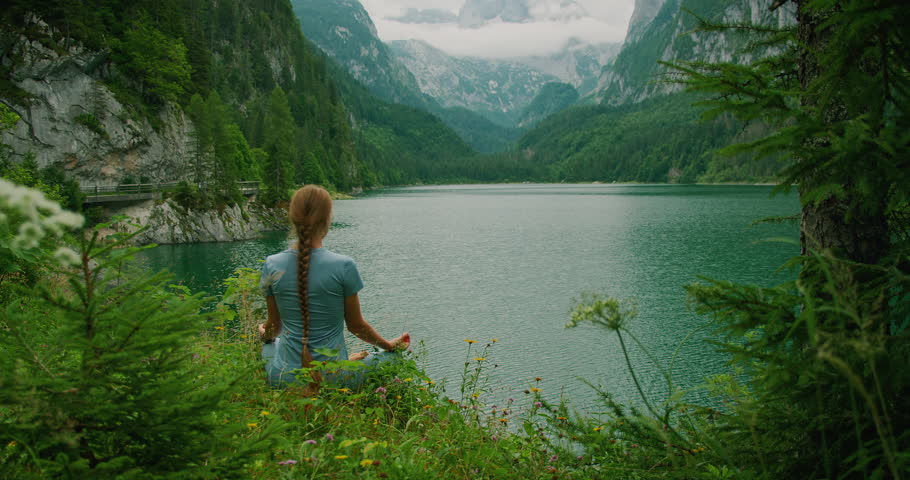 Woman sits on a grassy lakeshore, meditating with a mountain view at Gosau Lake in Austria
