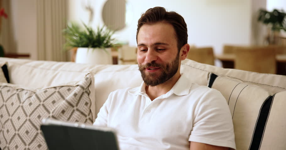 Tablet, video call and man on sofa in home for communication, connectivity or contact. Laugh, digital technology and male person talking with virtual conversation on app in living room at apartment.