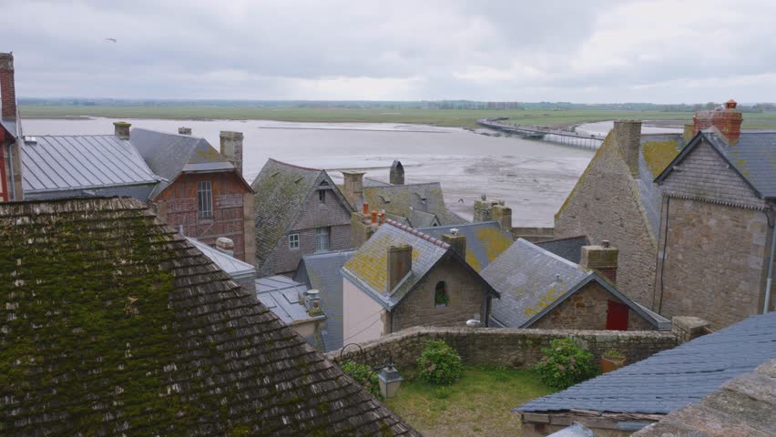 View from the Mont-Saint-Michel Abbey walls over the tiled rooftops of the old buildings, new bridge and the English Channel at low tide on a windy spring day.