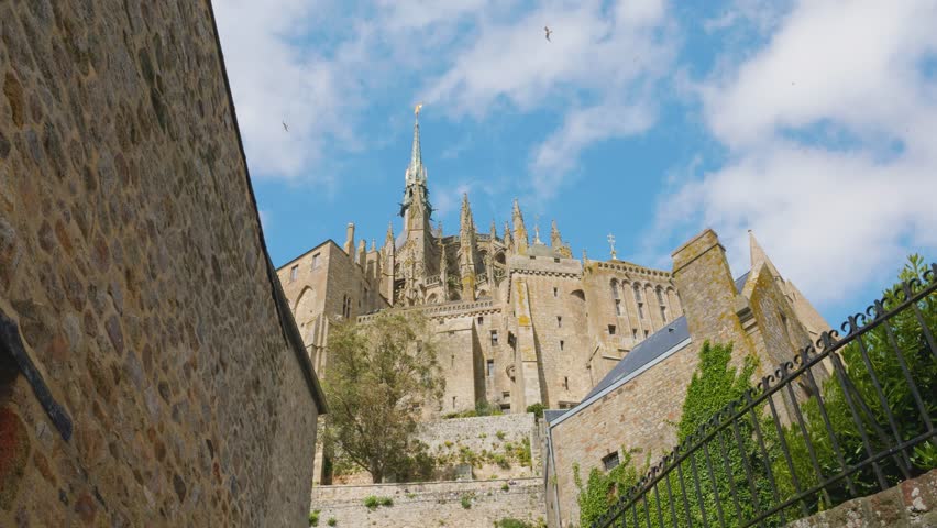 View of the Mont-Saint-Michel Abbey walls and soaring seagulls against a blue sky and clouds on a windy spring day.