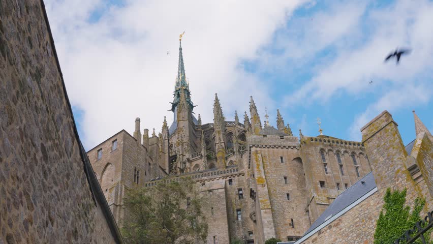 View of the Mont-Saint-Michel Abbey walls and soaring seagulls against a blue sky and clouds on a windy spring day.