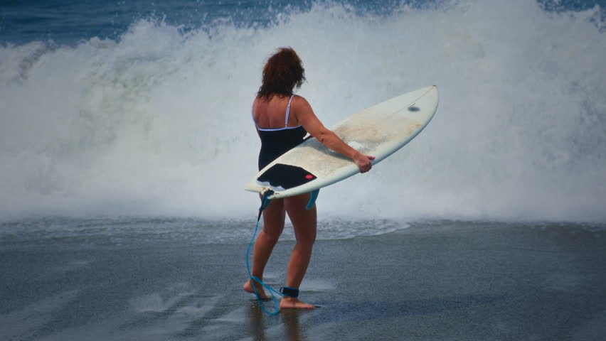 Old surfer woman walks along a Bali sandy beach, holding or carrying a surfboard. Senior female walks to the ocean or sea for surfing training or competition, waiting for waves. Surf session