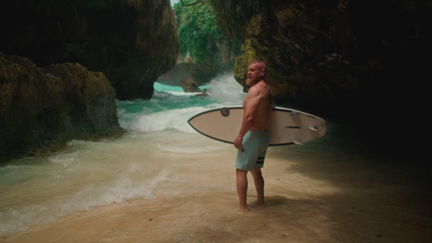 Sporty old man holds a surfboard on seashore. Senior male surfs in Bali, surfing on Uluwatu beach. Travelling elderly people enjoying retired lifestyle on tropical island vacation trip. Portrait