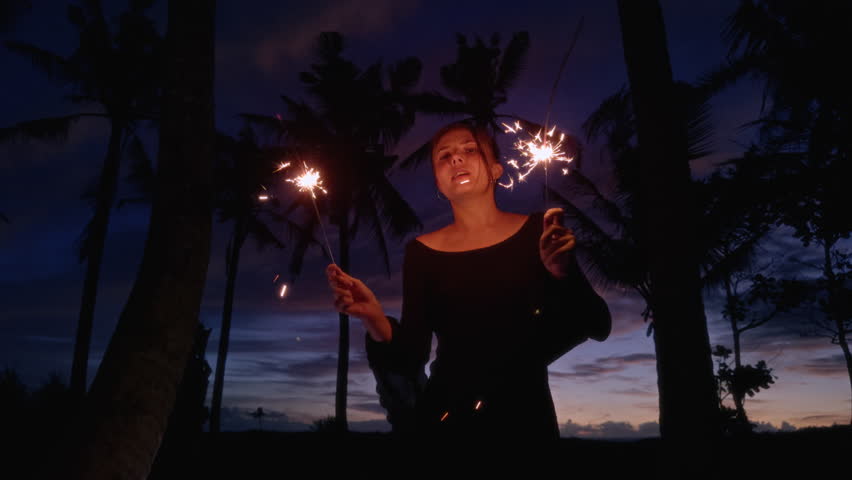 Young woman celebrates the new year with sparklers or Bengali fire. Beautiful girl is dancing under palm tree on a beach in a tropical country. Winter vacation in Bali. Female, Christmas light, x-mas