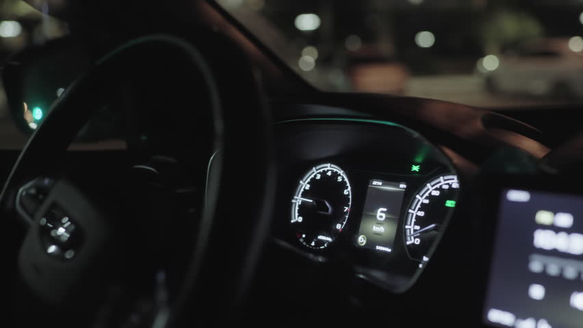 Close up of driver turning steering wheel inside moving car at night with illuminated dashboard, digital speedometer, and blurred street lights visible through windshield