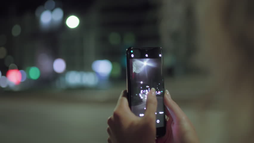 Close up of woman using smartphone camera to take photo of illuminated street at night, fingers adjusting focus on screen, colorful city lights in background