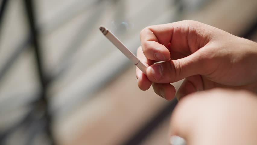close up hand view of smoker gently flicking cigarette as ash falls with subtle smoke trail against softly blurred outdoor setting in daylight