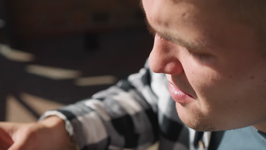 Close up of young man smoking almost finished cigarette while exhaling smoke under bright sunlight with blurred urban structure in background