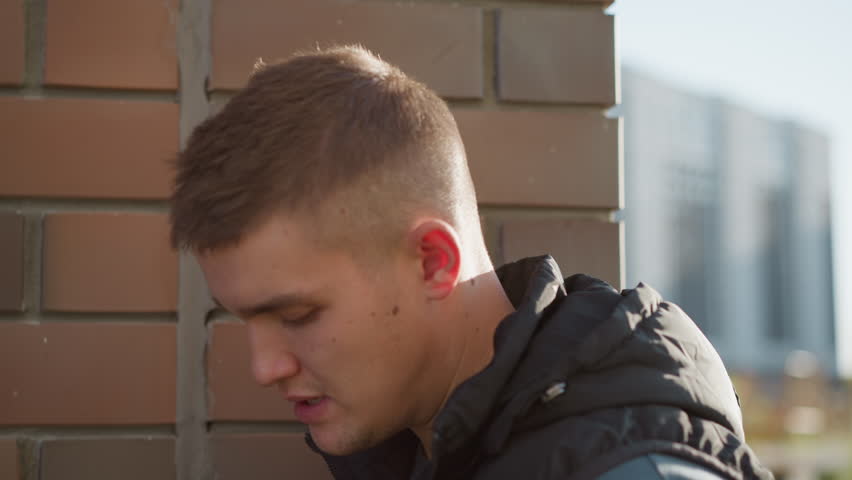 boy leaning close to brick wall secretly inhaling shisha and beginning to cough with blur view of distant office building in background under natural daylight wearing black vest and dark shirt