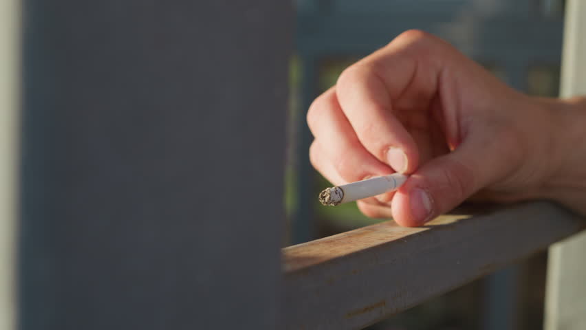 close up of fair skinned person casually holding cigarette over metal surface while wind blows ash and fingers slowly roll cigarette under soft daylight in relaxed urban setting