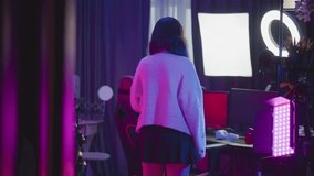 Young girl in fluffy sweater walking into neon lit studio adjusts seat at desk with ring light and white headphone then sits before computer monitors and LED panels under pink purple ambient glow - Powered by Shutterstock - Get 15% off with code: PIKWIZARD15