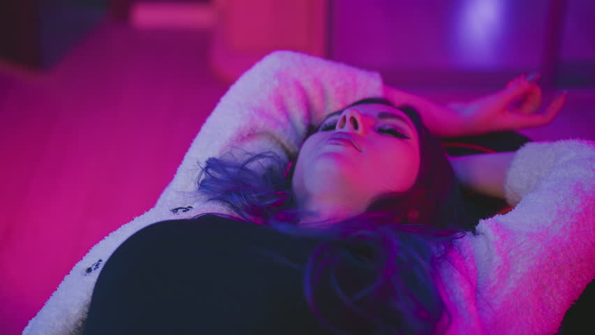 Young woman lying flat asleep with arms over head on soft surface, illuminated by vibrant pink and purple lighting, creating peaceful yet artistic indoor atmosphere with dreamy cinematic mood